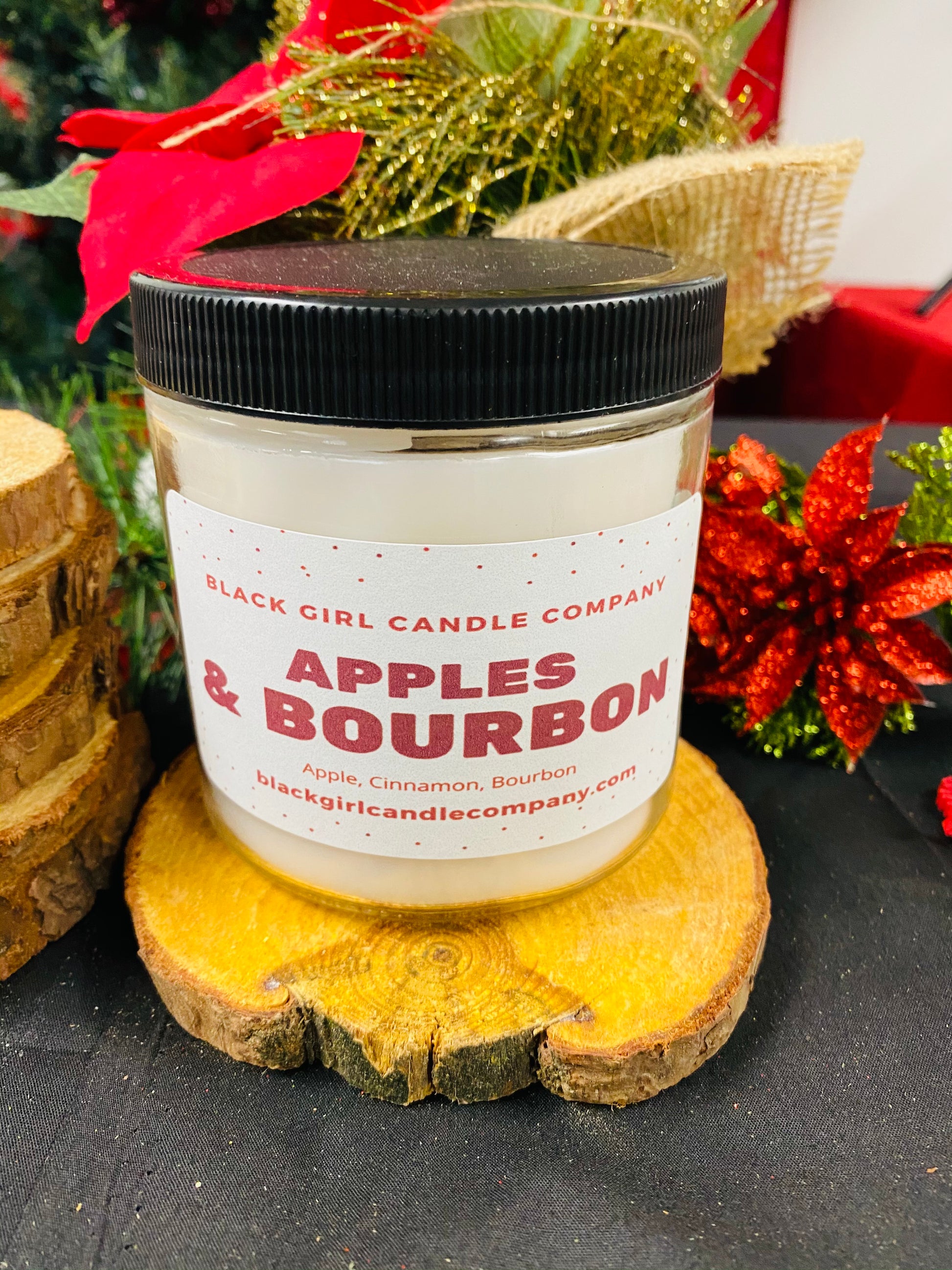 Black Girl Candle Company "Apples & Bourbon" Candle. White wax in clear jar with white label and black lid. Resting on wooden coaster, red and gold flowers in background